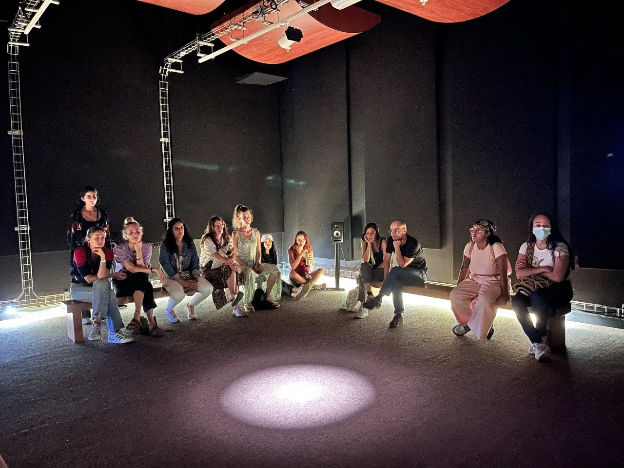Un grupo de oyentes sentados alrededor de los bordes de una habitación oscura con altavoces en trozos de metal, un círculo de luz en el suelo.